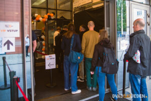 Ľudia stoja v rade pri vstupe označenom VIP VSTUP, s balónmi na ráme a smerovými značkami v modernej interiérovej hale.