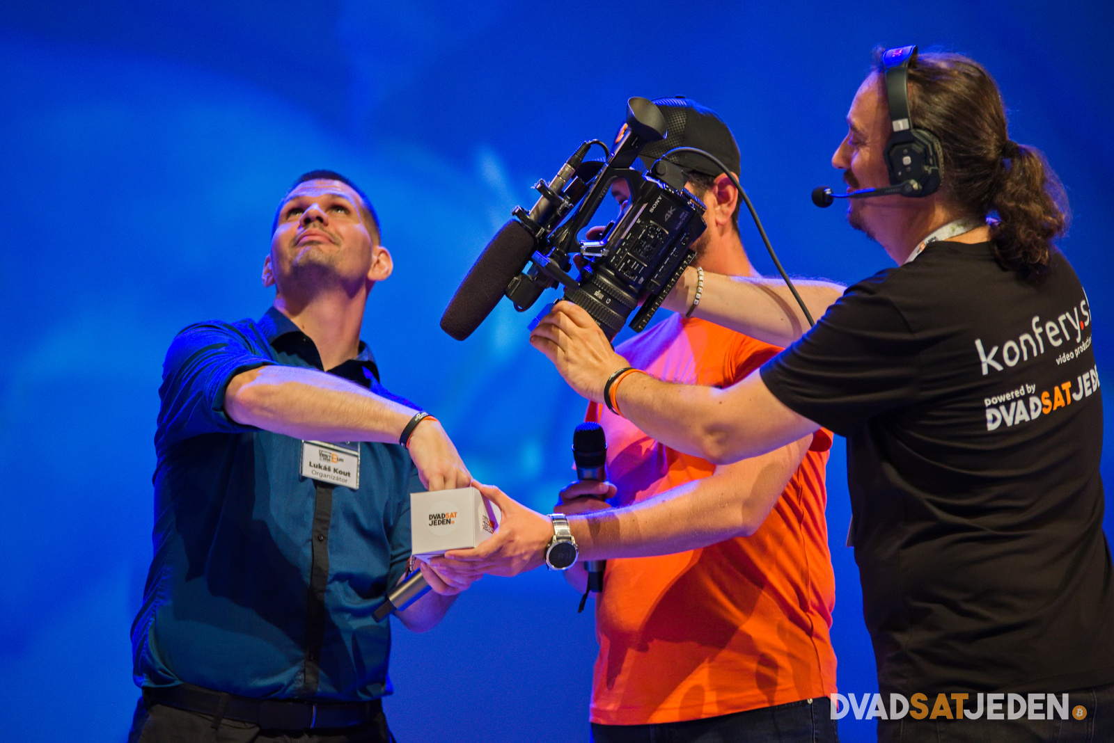 Traja muži stoja na pódiu; jeden ťahá papier z krabice, druhý drží kameru, tretí hovorí do mikrofónu. Modré pozadie.