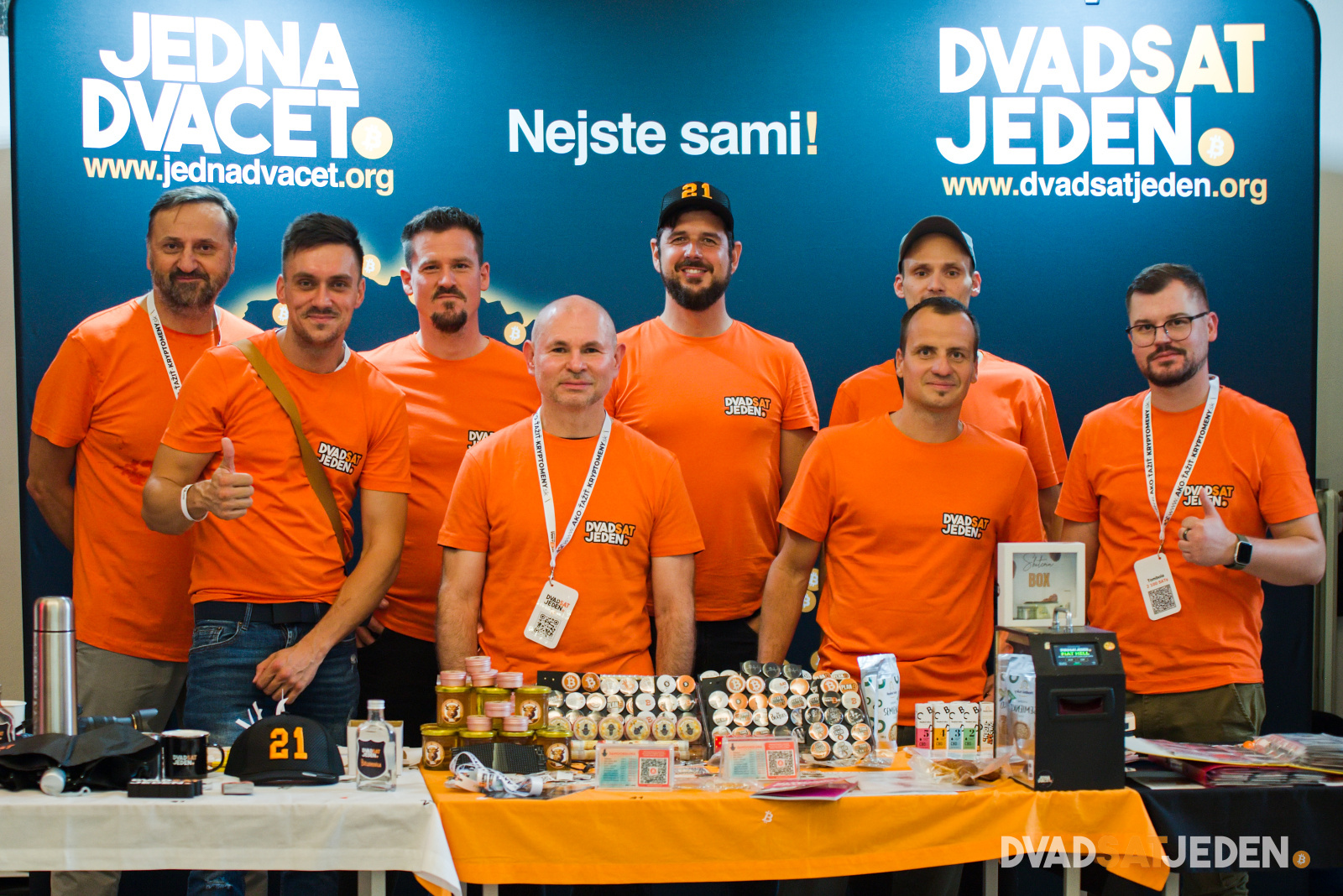 Osem mužov v oranžových tričkách stojí za stolom s tovarom a materiálmi na stánku s nápisom Jedna Dvacet a Dvadsať Jedna.