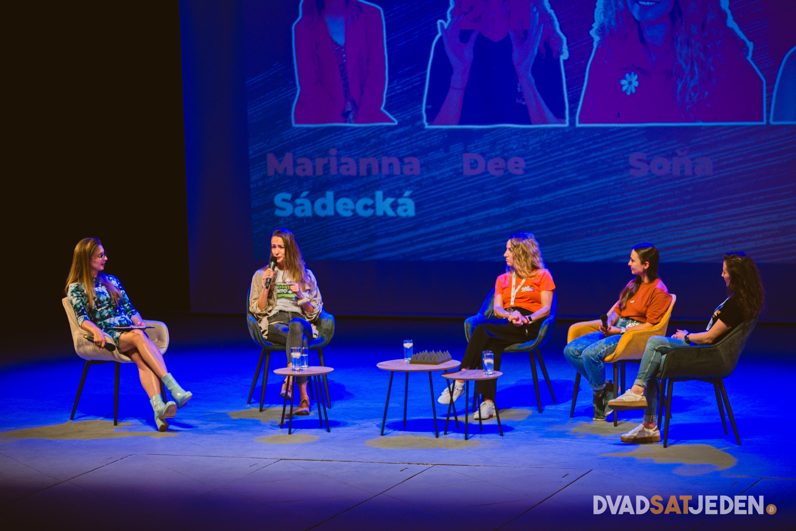 Päť ľudí sedí na pódiu na stoličkách a diskutuje. Za nimi sú premietnuté mená, vpredu je logo "DVADSATJEDEN".