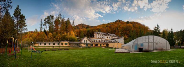 Na trávnatej ploche pred veľkou budovou je detské ihrisko a skleník, v pozadí sú lesnaté kopce a čiastočne oblačná obloha.