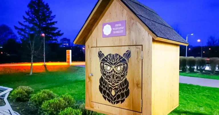 Drevená konštrukcia s ilustráciou sovy na dverách stojí na tráve pri chodníku za súmraku, v pozadí sú stromy a lampy.