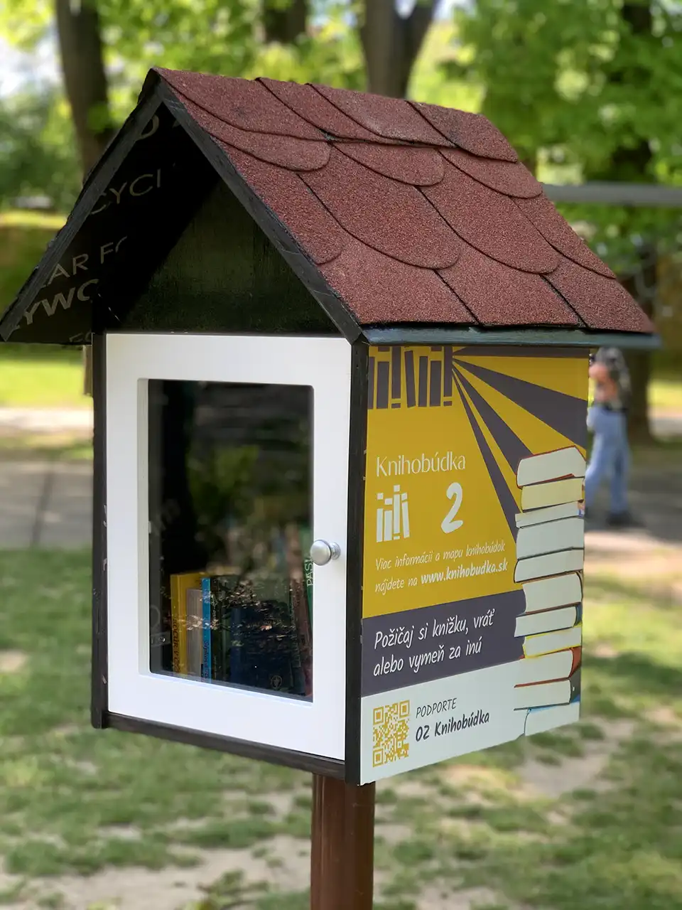 Malá vonkajšia knižná búdka so sklenenými dvierkami a slovenským nápisom v parku, v pozadí sú stromy.